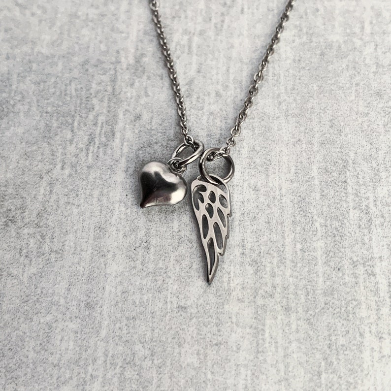 May include: A silver necklace with a heart and angel wing charm. The necklace is on a grey background.