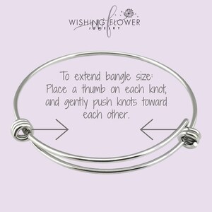 May include: A silver expandable bangle bracelet with two knots on either end. The bracelet is shown on a light purple background with text that reads: "To extend bangle size: Place a thumb on each knot, and gently push knots toward each other."