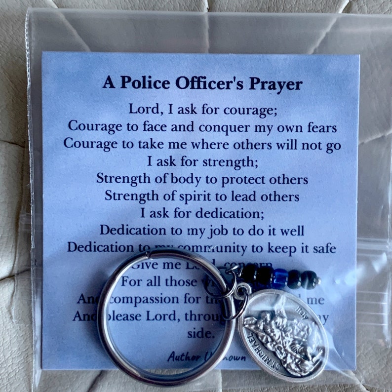 May include: A blue card with a prayer for police officers. The card reads "A Police Officer's Prayer" and includes a prayer asking for courage, strength, and dedication. The card is packaged with a silver key chain featuring a St. Michael medal.