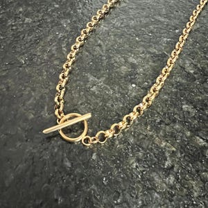 May include: A gold-colored necklace with a toggle clasp. The chain is made of rounded links. The toggle clasp is a bar that fits through a circular loop. The necklace is displayed on a dark, textured surface.