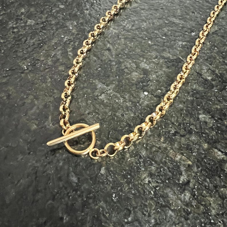 May include: A gold-colored necklace with a toggle clasp. The chain is made of rounded links. The toggle clasp is a bar that fits through a circular loop. The necklace is displayed on a dark, textured surface.