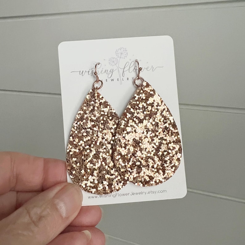 May include: A pair of rose gold glitter teardrop earrings with a rose gold hook fastening. The earrings are displayed on a white card with the text "Wishing Flower Jewelry" and a website address.