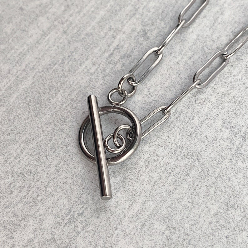 May include: A silver chain necklace with a toggle clasp. The clasp is a round ring with a bar that slides through it.