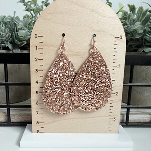 May include: A pair of rose gold glitter teardrop earrings. The earrings are approximately 5 cm long and are shown on a wooden display stand with a ruler.