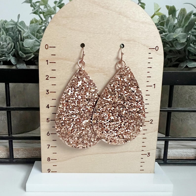 May include: A pair of rose gold glitter teardrop earrings. The earrings are approximately 5 cm long and are shown on a wooden display stand with a ruler.