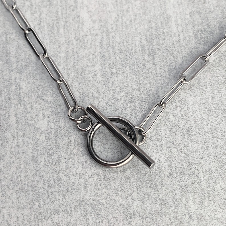 May include: A silver chain necklace with a toggle clasp. The clasp is a round ring with a straight bar that slides through it.