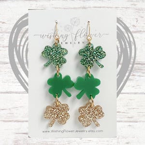 May include: A pair of dangle earrings featuring three shamrock charms. The top charm is green glitter, the middle charm is solid green, and the bottom charm is gold glitter. The earrings are on a white card with the text "Wishing Flower Jewelry" and a website address.