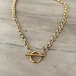 May include: A gold-colored necklace with a toggle clasp. The chain is made of small, rounded links. The clasp features a circular ring and a straight bar. The necklace is displayed on a light gray wooden surface.