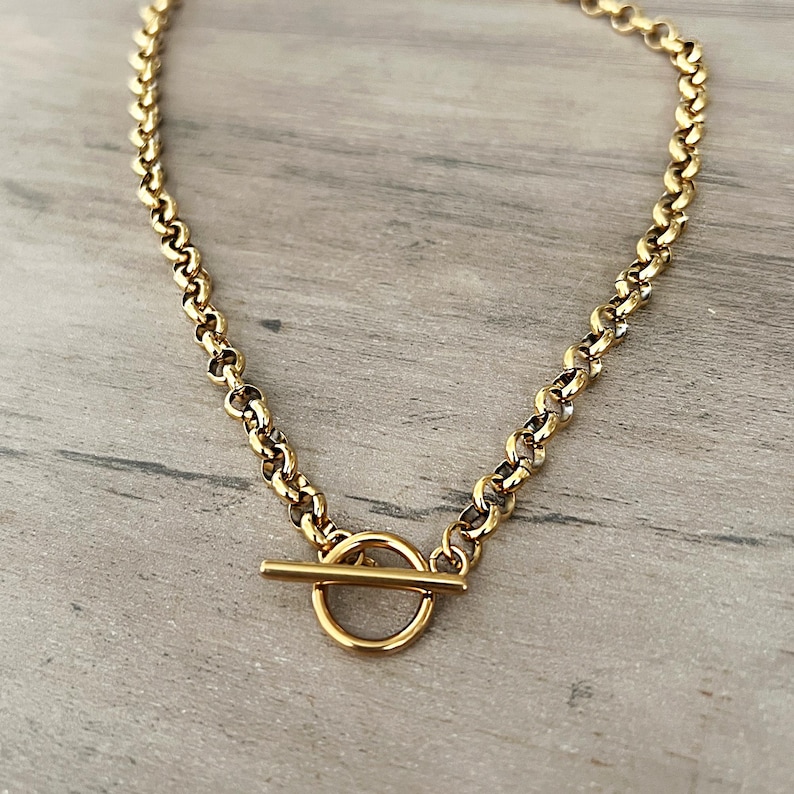 May include: A gold-colored necklace with a toggle clasp. The chain is made of small, rounded links. The clasp features a circular ring and a straight bar. The necklace is displayed on a light gray wooden surface.