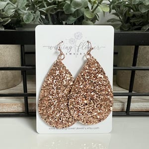 May include: Rose gold glitter teardrop earrings with a silver hook. The earrings are on a white card with the text "Wishing Flower Jewellery" and a website address.