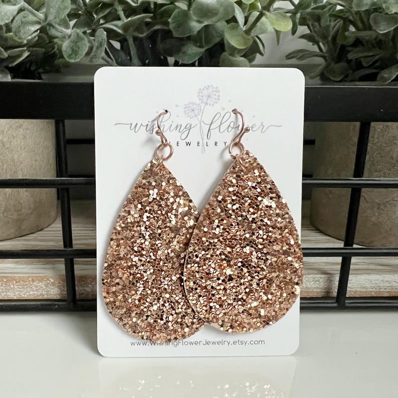 May include: Rose gold glitter teardrop earrings with a silver hook. The earrings are on a white card with the text "Wishing Flower Jewellery" and a website address.