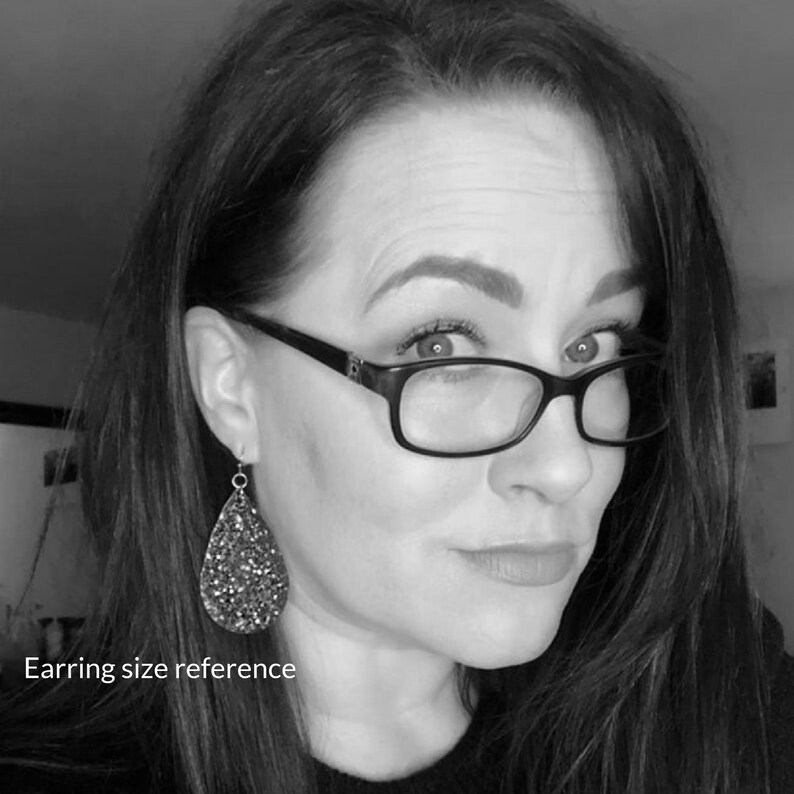 May include: A woman wearing black framed glasses with a teardrop-shaped glitter earring. The earring is silver and has a sparkly, iridescent finish.  Earring size reference.