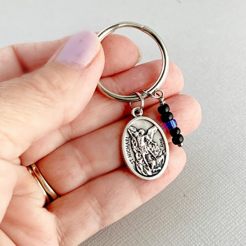 May include: Silver keychain with a Saint Michael medal and black and blue beads. The medal features a depiction of Saint Michael slaying a dragon. The text "St. Michael" and "Italy" are inscribed on the medal.