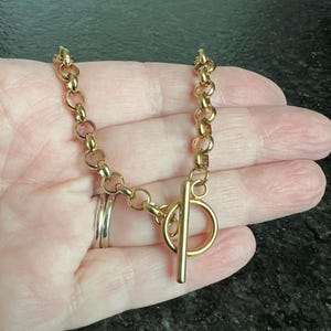 May include: A gold-colored chain bracelet with a toggle clasp. The bracelet features a series of rounded links connected together. The toggle clasp has a circular ring and a straight bar. The bracelet is shown on a hand.