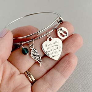 May include: Silver bangle bracelet with charms, including a heart charm with the inscription "I will hold you in my heart until I hold you in heaven", a small green bead, and a wing charm.