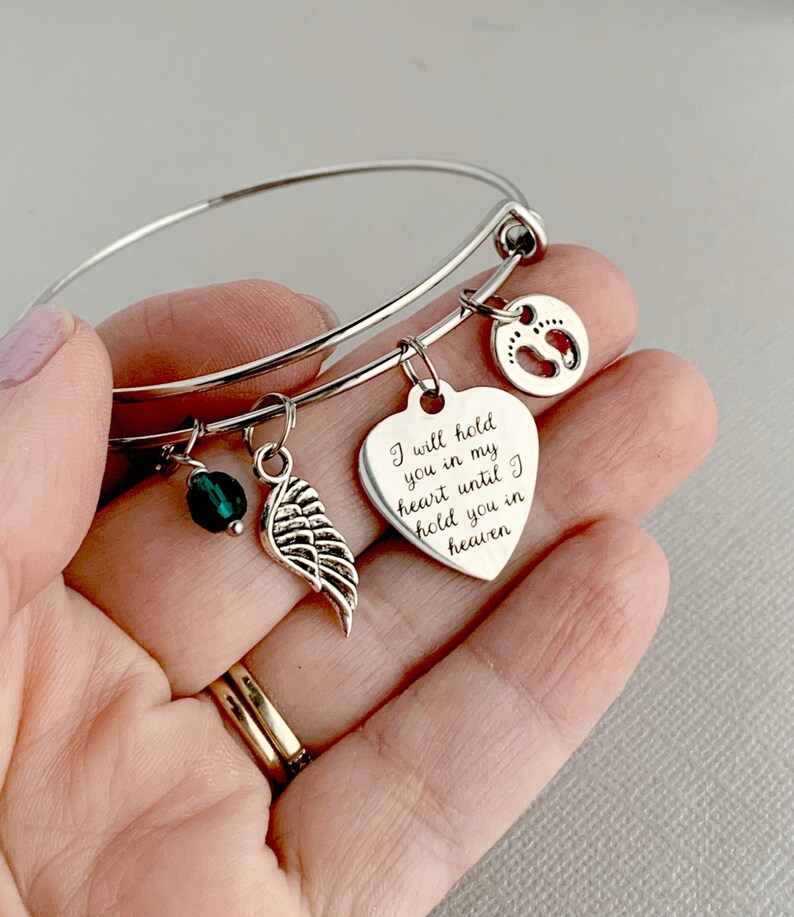 May include: Silver bangle bracelet with charms, including a heart charm with the inscription "I will hold you in my heart until I hold you in heaven", a small green bead, and a wing charm.