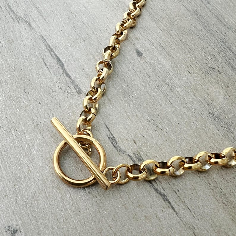 May include: Gold-toned necklace with a toggle clasp. The chain features a series of rounded links. The clasp consists of a circular ring and a straight bar. The jewelry is displayed on a gray surface.
