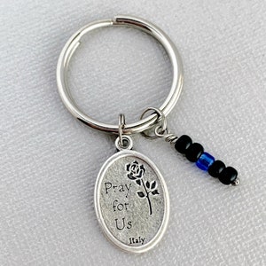 May include: Silver key chain with a silver oval charm that says "Pray for Us" with a rose design and "Italy" at the bottom. The charm is attached to a silver key ring with a black and blue beaded chain.