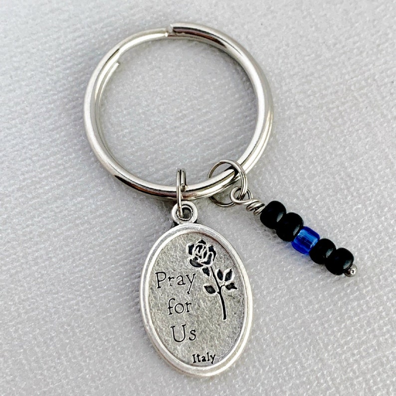 May include: Silver key chain with a silver oval charm that says "Pray for Us" with a rose design and "Italy" at the bottom. The charm is attached to a silver key ring with a black and blue beaded chain.