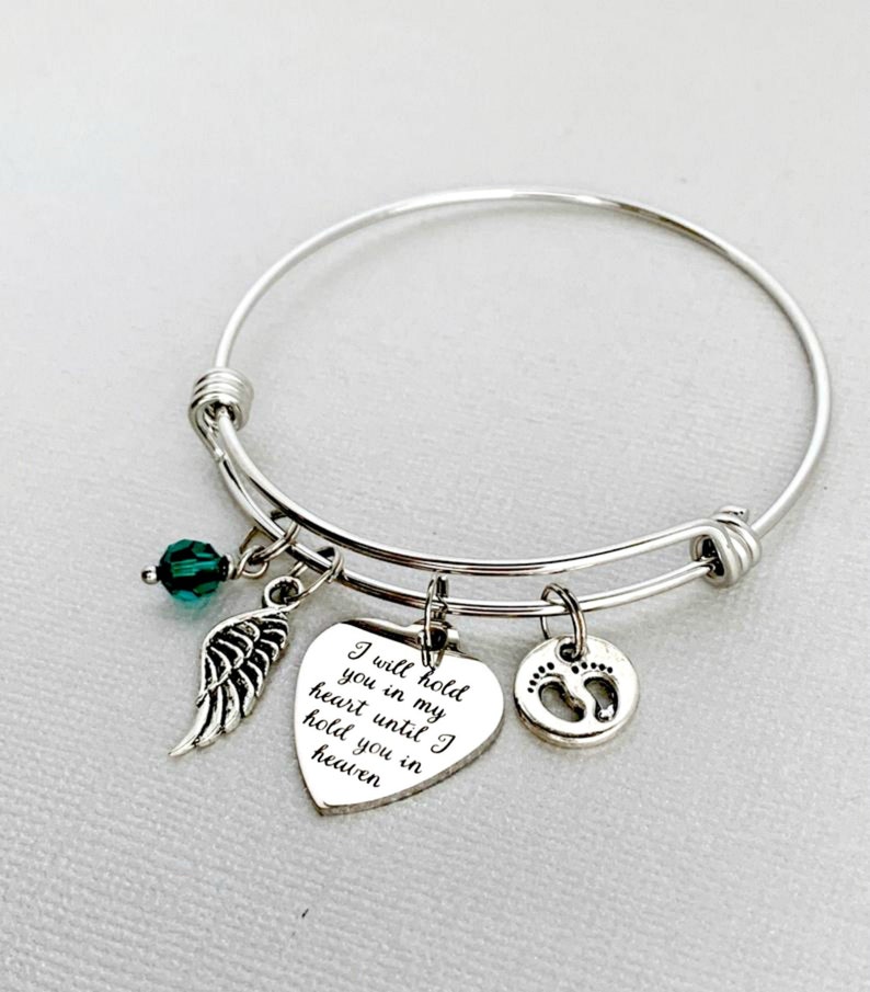 May include: A silver bangle bracelet with a heart charm that reads 'I will hold you in my heart until I hold you in heaven.' The bracelet also has a small green crystal charm, a silver angel wing charm, and a silver charm with a footprint design.