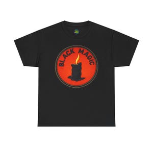 May include: A black short-sleeve t-shirt featuring a circular graphic on the chest. The graphic has a bright orange background with a black candle and a yellow flame in the center. The text BLACK MAGIC is printed in black around the candle.