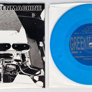 May include: A blue vinyl record and album cover for "GREENMACHINE." The album cover is black and white, featuring an engine. The record is a vibrant blue, with the band's name and song titles printed on it. A classic music collectible.