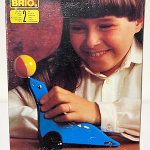 May include: A blue wooden toy seal with a red and yellow ball on its nose. The seal is on a brown surface. The toy is for children ages 2 and up. The box has the text "BRIO" and "From 2 years".