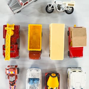 May include: Collection of vintage toy vehicles, including a silver car, white police motorcycle, red fire truck, yellow dump truck, beige bus, and a red and tan truck. Also included are a race car, white boat, cement truck, and a white van.