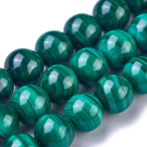 Peut inclure: Gros plan sur un fil de perles de malachite polies. Les perles sont d'un vert profond avec des motifs tourbillonnants, reflétant la lumière. Les perles rondes sont enfilées ensemble, mettant en valeur la beauté naturelle de la pierre.
