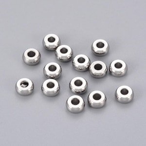 Peut inclure: Un ensemble de perles métalliques argentées et brillantes. Chaque perle est ronde avec un trou traversant le centre. Les perles sont dispersées sur une surface gris clair, reflétant la lumière.