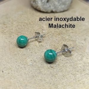 Peut inclure: Boucles d'oreilles à clous avec des pierres rondes de malachite verte montées sur des tiges en acier inoxydable. Finition polie. Le texte "acier inoxydable Malachite" est visible. Présentées sur une surface brun clair.