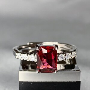 Peut inclure: Une bague en argent avec une grande pierre rouge de forme carrée entourée de petites pierres claires. La bague est présentée sur une boîte noire avec le mot "etc." imprimé en blanc.