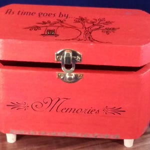 May include: A red wooden memory box with a hinged lid and a silver clasp. The lid is decorated with a tree and a birdhouse, and the box has the words "As time goes by..." and "Memories" written on it.