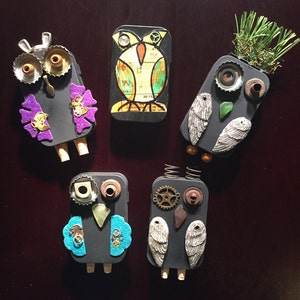 May include: Five handmade owl ornaments made from recycled materials. Each owl has a different design and color scheme, including purple, blue, green, and brown. The owls are decorated with various embellishments, such as buttons, beads, and metal pieces.