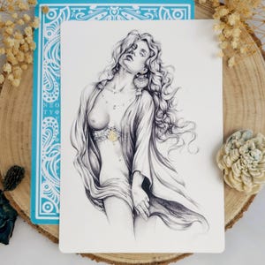 Puede incluir: Un dibujo a lápiz en blanco y negro de una mujer con cabello largo y suelto y una prenda drapeada. La obra de arte está sobre una tarjeta rectangular blanca, con una tarjeta azul detrás. La tarjeta azul tiene patrones decorativos blancos y texto. La obra de arte está colocada sobre una rodaja de madera.