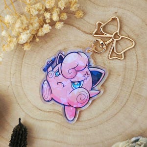 Könnte beinhalten: Ein rosa-blaues Cartoon-Charakter-Schlüsselanhänger mit einem goldfarbenen, schleifenförmigen Verschluss. Der Charakter hat einen wütenden Gesichtsausdruck mit blauen Augen und einem Kreuz. Der Schlüsselanhänger liegt auf einer Holzoberfläche.