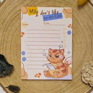 Könnte beinhalten: Notizblock mit dem Text "My, I don't like to-do-list" und einer Cartoon-Katzen-Illustration. Die Katze ist orange und weiß, sieht verärgert aus, während sie ein Papier hält. Der Notizblock hat einen linierten Abschnitt zum Schreiben.