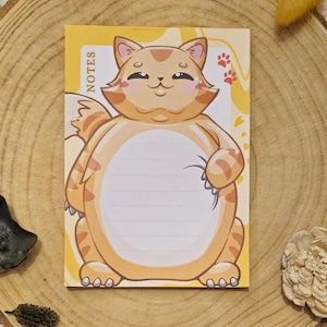 Könnte beinhalten: Ein gelber Notizblock mit einer Cartoon-Katzen-Illustration. Die Katze ist orange und weiß mit einem lächelnden Gesicht und einem großen Bauch. Der Notizblock hat linierte Seiten und das Wort "NOTES" vertikal auf der Seite gedruckt.