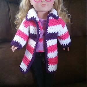 May include: A doll wearing a pink and purple striped crocheted cardigan with a white collar. The doll has blonde hair and is wearing pink glasses and pink shoes.