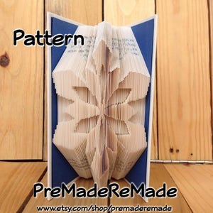 May include: A white book folded into a 3D snowflake shape. The book is open and the pages are folded to create a detailed snowflake design. The book is sitting on a blue background. The text "Pattern" is at the top of the image and the text "PreMadeReMade" is at the bottom of the image.