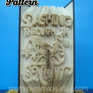 May include: A book art sculpture with the words "Sunshine Through the Storm" folded into the pages. The book is closed and standing upright on a brown surface.
