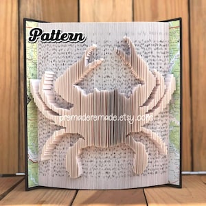 Op de afbeelding: Een boek gevouwen in de vorm van een krab. Het boek is gemaakt van wit papier met zwarte tekst. De krab is lichtbruin. Het boek heeft de titel "Pattern" en heeft de website "premadehandmade.etsy.com" erop gedrukt.