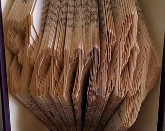 May include: A book art sculpture, folded into the shape of the word "LOVE", with the word "Pattern" written above it. The book is open and the pages are folded into a three-dimensional shape. The book is bound in a purple cover and has a purple lace trim at the bottom.