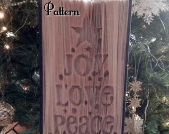 May include: A book with the words "Joy, Love, Peace, Believe" carved into the pages, forming a Christmas tree shape. The book is standing upright on a wooden surface. The text "Pattern" is visible at the top of the book. The text "Pre Made Re Made 2015" is visible at the bottom of the book.