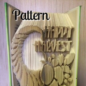 Puede incluir: Un libro con las palabras "Happy Harvest" dobladas en las páginas para crear un diseño 3D. El libro está abierto y las páginas están dobladas para crear un diseño de girasol. El libro está sobre una superficie de madera.