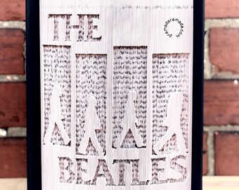 May include: A handmade book sculpture featuring the words "THE BEATLES" cut into the pages. The design includes silhouettes of the band members. The book has a black cover and is displayed against a brick wall.