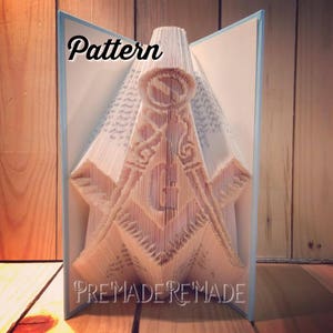 May include: A book folded into a 3D sculpture of a Masonic symbol. The sculpture is made of white paper and is standing on a wooden surface. The text "Pattern" is written at the top of the sculpture and the text "PreMadeReMade" is written at the bottom.