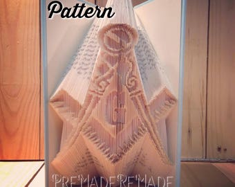 May include: A book folded into a 3D sculpture of a Masonic symbol. The sculpture is made of white paper and is standing on a wooden surface. The text "Pattern" is written at the top of the sculpture and the text "PreMadeReMade" is written at the bottom.