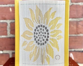 May include: A folded book art sculpture featuring a sunflower design created by precisely folding the pages. The book has bright yellow covers and the sunflower pattern is highlighted in yellow against the white paper edges of the pages.