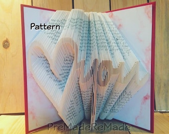 May include: A book folded into the shape of a heart, with the word "Pattern" written on the cover. The book is open and the pages are folded to create a three-dimensional heart shape. The book is on a wooden surface.